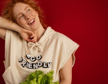 Cheerful woman with red hair in eco-friendly attire holding fresh lettuce against a vibrant red background.