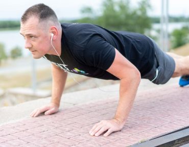 Adult male exercising outdoors performing push-ups with earphones and water bottle.