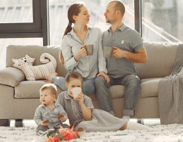 Happy family with children spending time at home while parents sitting on couch with cups of coffee and looking at each other when kids resting on floor