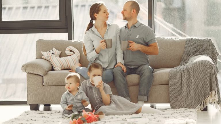 Happy family with children spending time at home while parents sitting on couch with cups of coffee and looking at each other when kids resting on floor